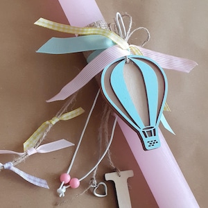 Può includere: Una candela pasquale rosa decorata con nastri di colore azzurro pastello, giallo e rosa, una mongolfiera di legno e una lettera I di legno.