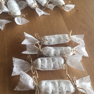 Puede incluir: Bolsas de regalo de encaje blanco atadas con cuerda y llenas de caramelos blancos. Las bolsas est&aacute;n dispuestas sobre una superficie de madera.