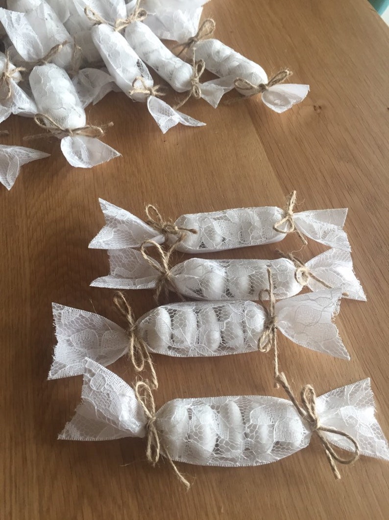 Puede incluir: Bolsas de regalo de encaje blanco atadas con cuerda y llenas de caramelos blancos. Las bolsas est&aacute;n dispuestas sobre una superficie de madera.