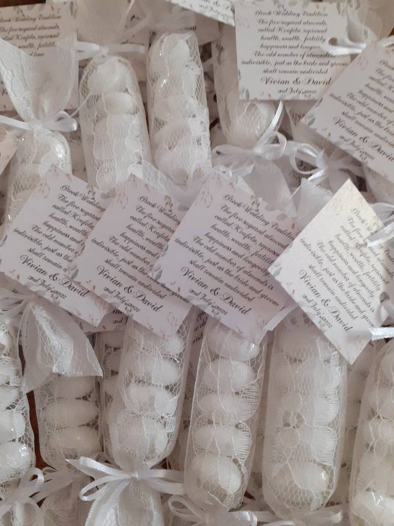 Puede incluir: Bolsitas de encaje blanco llenas de almendras blancas. Cada bolsa tiene una peque&ntilde;a tarjeta adjunta con el texto "Tradici&oacute;n griega de boda. Las cinco almendras azucaradas llamadas Koufeta, representan la salud, la riqueza, la fertilidad, la felicidad y la longevidad. El n&uacute;mero impar de almendras es indivisible, al igual que la novia y el novio permanecer&aacute;n indivisibles. Vivian & David 2 de julio de 2022."