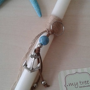 Può includere: Candela bianca con un avvolgimento di spago marrone e un ciondolo a forma di ancora argentata. Il ciondolo è attaccato a un cordino di cuoio marrone con una perla blu e perline argentate. Sullo sfondo c'è una stella marina. La carta recita "my tree handmade" con il sito web e le informazioni di contatto.