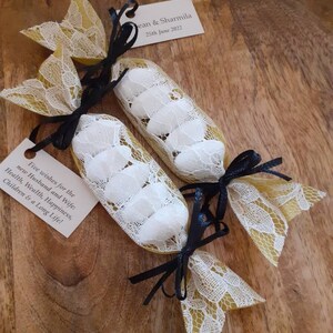 Puede incluir: Dos peque&ntilde;as bolsas de regalo de encaje blanco atadas con una cinta negra, llenas de almendras blancas. Las bolsas est&aacute;n sobre una superficie de madera y tienen una peque&ntilde;a tarjeta adjunta con el texto "Cinco deseos para los nuevos esposos: Salud, Riqueza, Felicidad, Hijos y Larga Vida!" y "Jean & Sharmila 25 de junio de 2022."