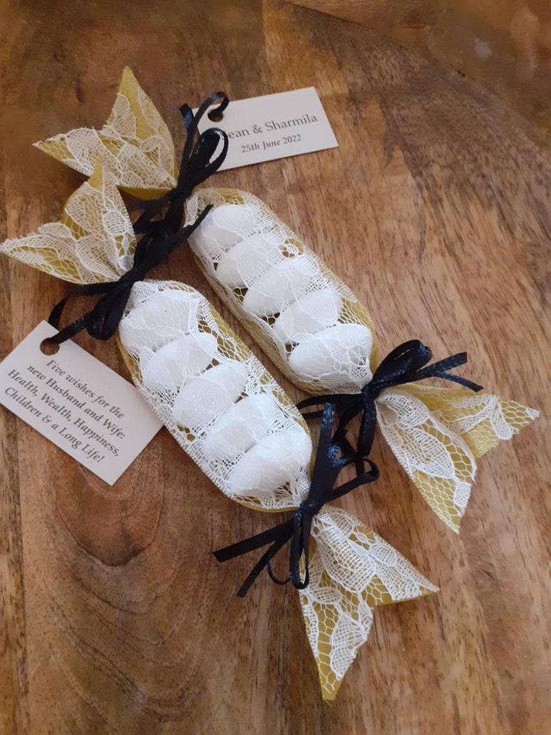 Puede incluir: Dos peque&ntilde;as bolsas de regalo de encaje blanco atadas con una cinta negra, llenas de almendras blancas. Las bolsas est&aacute;n sobre una superficie de madera y tienen una peque&ntilde;a tarjeta adjunta con el texto "Cinco deseos para los nuevos esposos: Salud, Riqueza, Felicidad, Hijos y Larga Vida!" y "Jean & Sharmila 25 de junio de 2022."