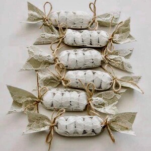 Puede incluir: Un conjunto de ocho peque&ntilde;os caramelos redondos blancos envueltos en encaje beige y atados con cuerda. Los caramelos est&aacute;n dispuestos en fila, con los extremos del envoltorio de encaje extendi&eacute;ndose hacia los lados.
