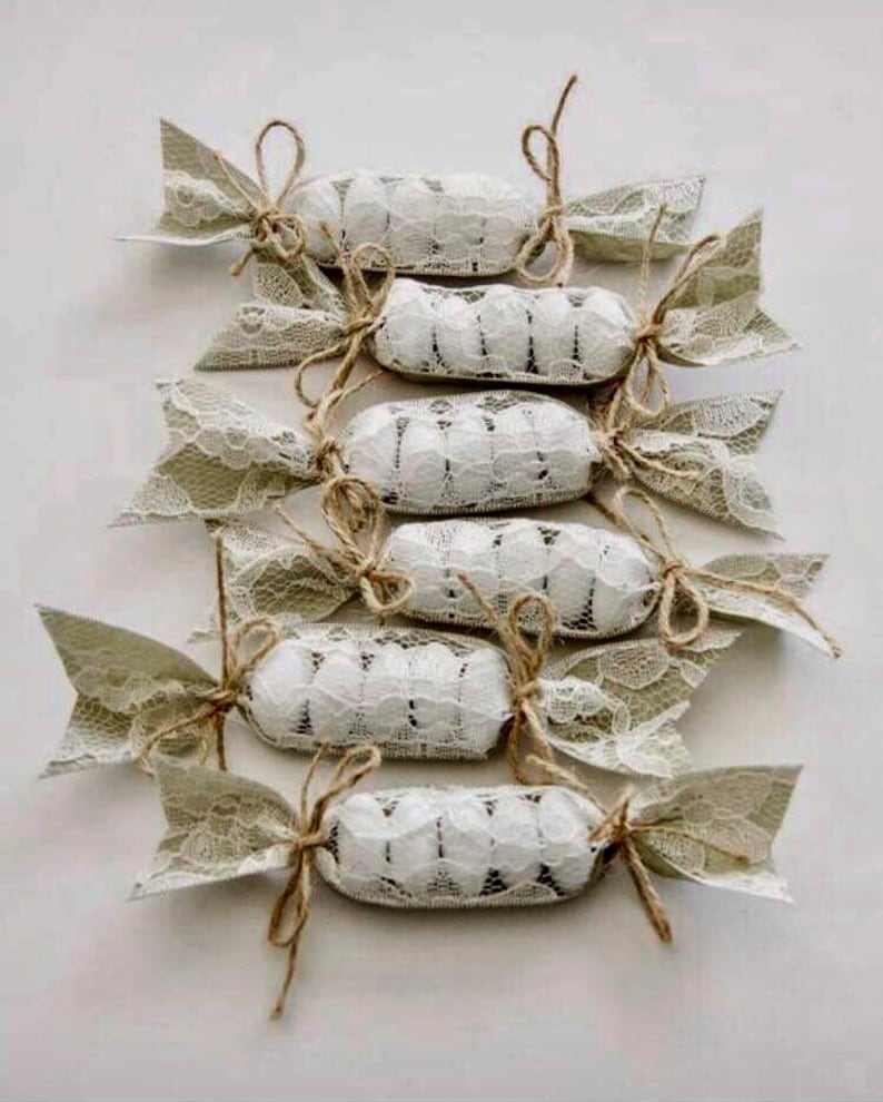Puede incluir: Un conjunto de ocho peque&ntilde;os caramelos redondos blancos envueltos en encaje beige y atados con cuerda. Los caramelos est&aacute;n dispuestos en fila, con los extremos del envoltorio de encaje extendi&eacute;ndose hacia los lados.