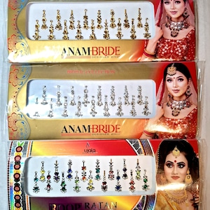 Op de afbeelding: Drie pakketten met decoratieve bindi-stickers. Het bovenste pakket heeft gouden bindi-stickers met de tekst "ANAMBRIDE DESIGNER BINDI". Het middelste pakket heeft zilveren bindi-stickers met de tekst "ANAMBRIDE DESIGNER BINDI". Het onderste pakket heeft kleurrijke bindi-stickers met de tekst "ROOP RATAN FANCY BINDI".