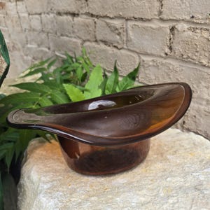 May include: A translucent amber-colored glass bowl shaped like an inverted top hat. The object features a wide, curved brim and a deep, rounded base, crafted from smooth, polished glass that allows light to pass through its warm, brownish hue.