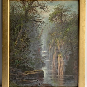 May include: An oil painting of a waterfall cascading through a lush, green forest. The artwork is framed in a gold-colored wooden frame. The painting depicts a serene landscape with detailed trees and rocks.