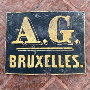 May include: A black metal sign with gold lettering. The sign reads "A.G. BRUXELLES."