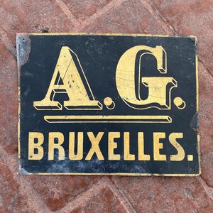 May include: A black metal sign with gold lettering that reads "A.G. BRUXELLES".