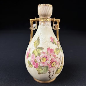 May include: A small, cream-colored porcelain vase with gold accents and a floral design. The vase has two handles and a narrow neck. The flowers are pink and white with green leaves.