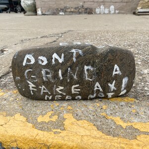 May include: A grey rock with the words "Don't Grind a Fake Axe!" written in white paint.