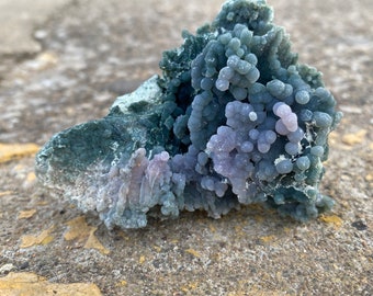 May include: A close-up of a purple and green agate geode with a rough, textured surface. The geode has a cluster of small, round nodules that are a light purple color. The nodules are surrounded by a darker green, almost black, agate.