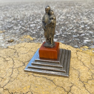 May include: A silver statue of a person holding a child, standing on a tiered, orange and silver base.