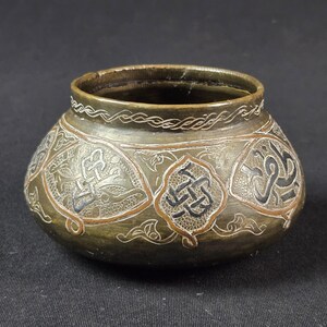 May include: A small, round, dark brass bowl featuring intricate hand-engraved patterns. The surface is decorated with alternating panels containing geometric knotwork and Arabic calligraphy, all highlighted with silver and copper-colored inlays.