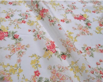 May include: White fabric with a repeating floral pattern. The design features pink, yellow, and peach-colored flowers with green stems and leaves. The fabric is folded, creating shadows and depth.