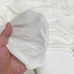 Pode incluir: Uma peça de tecido branco sujo com uma textura finamente canelada. O material é leve e ligeiramente translúcido, exibindo um delicado padrão de malha com linhas verticais que percorrem toda a superfície do tecido macio e flexível.