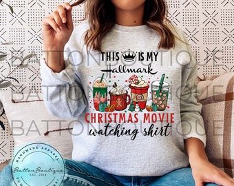 May include: A light gray crewneck sweatshirt featuring a graphic print with the text THIS IS MY Hallmark CHRISTMAS MOVIE watching shirt. The design includes illustrations of four festive holiday drinks, a small snowman, and wrapped gifts.