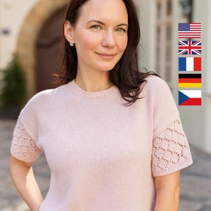 Könnte beinhalten: Ein kurzärmeliger Strickpullover in Hellrosa mit Rundhalsausschnitt und strukturierten Rauten-Lochmustern an den Ärmeln. Das Kleidungsstück ist auf der Vorderseite in glattem Rechtsstrick gefertigt und hat gerippte Abschlüsse.