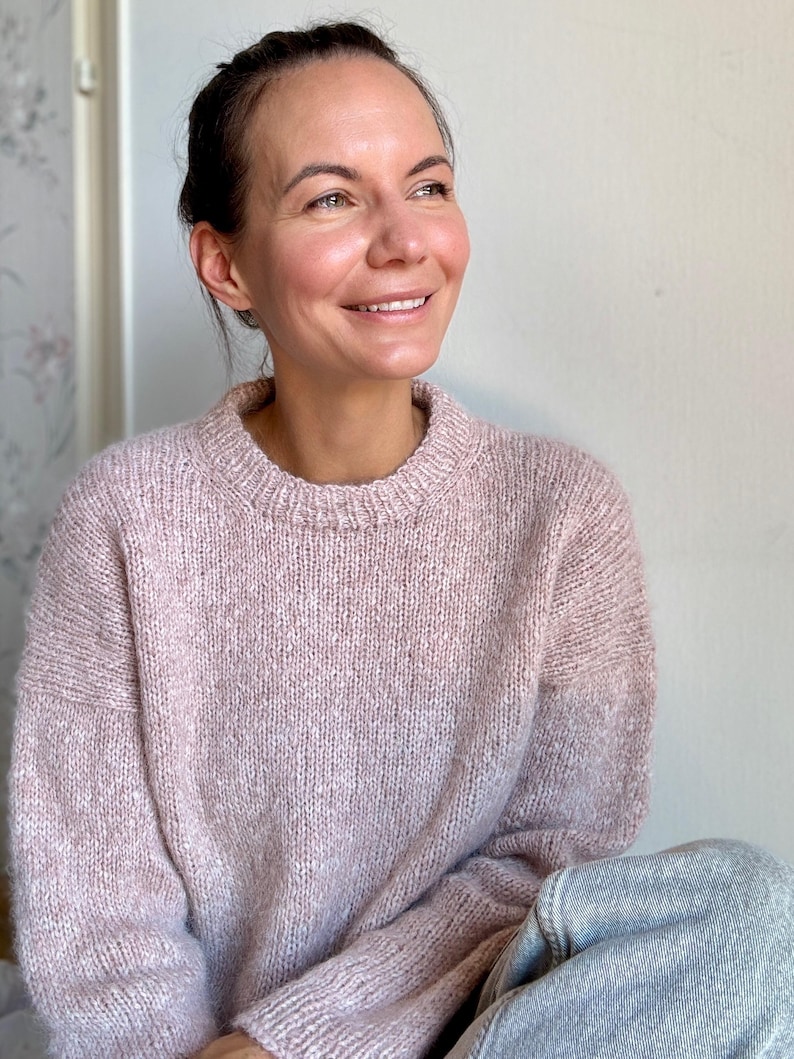 May include: A pale pink, oversized knit sweater with a relaxed fit and long sleeves. The sweater is made from a soft, fluffy yarn and has a simple, round neckline.  The sweater is shown draped over the lap of the model, showcasing its texture and drape. The light pink color is subtle and versatile, suitable for various casual outfits.