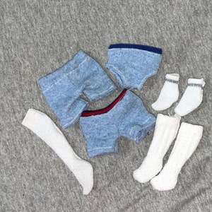 Può includere: Un set di indumenti blu e bianchi per una bambola, tra cui pantaloncini, una camicia e calzini. Gli indumenti sono realizzati in un tessuto morbido e confortevole.