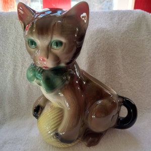 May include: A brown ceramic cat figurine with green eyes, a green bow, and a yellow yarn ball. The cat is sitting on its haunches with its tail curled around the yarn ball.