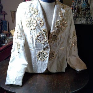 May include: A white silk jacket with gold and cream floral embroidery and sequins. The jacket has a button closure and long sleeves.