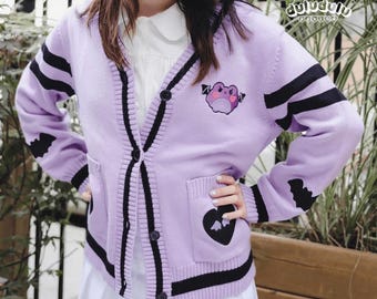 May include: A lilac cardigan with black stripes and trim. The cardigan features a frog and heart design. The sleeves have black bat-shaped accents. The cardigan has pockets and is buttoned up. The brand name "duludulu design" is visible.