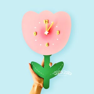 以下が含まれることがあります： 黄色い針と数字が付いた、ピンク色の花の形をした壁掛け時計。 時計には緑色の茎と葉があります。 時計は水色の背景に配置されています。 時計の針は12時を指しています。