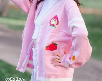 May include: Pale pink cardigan with white striped detailing on the sleeves and trim. The sweater has embroidered strawberry and frog motifs. A strawberry design is on the pocket. The brand name "duludulu" is at the bottom left.