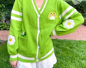 May include: A lime green cardigan with white stripes and daisy and frog embroidery. The V-neck sweater has white buttons and a pocket with a floral design. The sleeves have white stripes and a daisy patch.