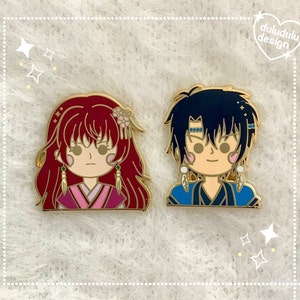 May include: Two enamel pins featuring anime-style characters with gold outlines. One pin has a red-haired character in pink and red clothing. The other pin has a black-haired character in blue clothing. The pins are on a white, textured surface.