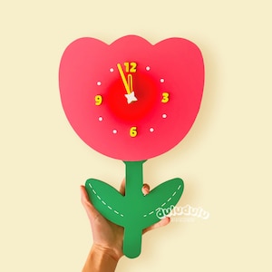 May include: A whimsical wall clock shaped like a red flower with a green stem and leaves. The clock face features yellow numbers and hands, with white dots marking the hours. The clock measures approximately 12 inches in height.