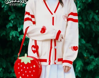 May include: Cream-colored cardigan with red stripes and heart and bear embroidery. A red strawberry-shaped crossbody bag is featured. The cardigan has a V-neck and button closure. The bag has a long strap.