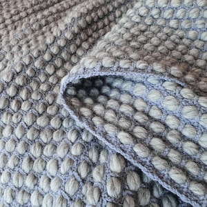Peut inclure: Gros plan sur une couverture grise textur&eacute;e avec un motif en relief en forme de bulles. La couverture a un bord pli&eacute; dans une nuance de gris plus claire, r&eacute;v&eacute;lant la texture et la construction du tissu tricot&eacute;. Th&egrave;me g&eacute;n&eacute;ral : d&eacute;coration int&eacute;rieure.