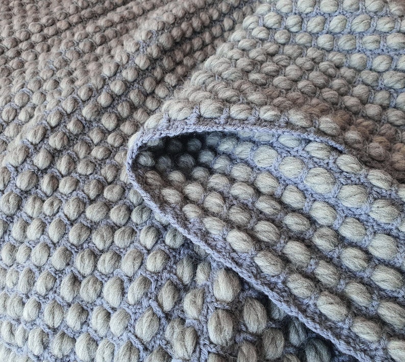 Peut inclure: Gros plan sur une couverture grise textur&eacute;e avec un motif en relief en forme de bulles. La couverture a un bord pli&eacute; dans une nuance de gris plus claire, r&eacute;v&eacute;lant la texture et la construction du tissu tricot&eacute;. Th&egrave;me g&eacute;n&eacute;ral : d&eacute;coration int&eacute;rieure.