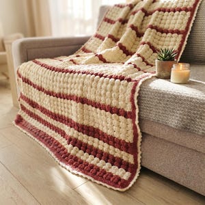 A crocheted throw blanket with a striped pattern in cream and burgundy. The blanket has a textured, puffy design.