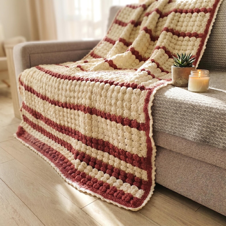 A crocheted throw blanket with a striped pattern in cream and burgundy. The blanket has a textured, puffy design.
