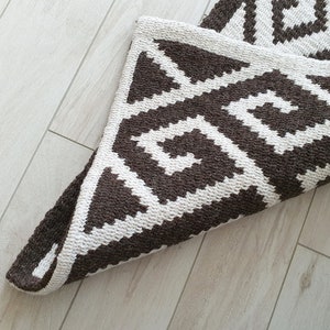 May include: A brown and white geometric patterned rug with a folded edge. The rug has a modern design with a repeating pattern of triangles and squares.