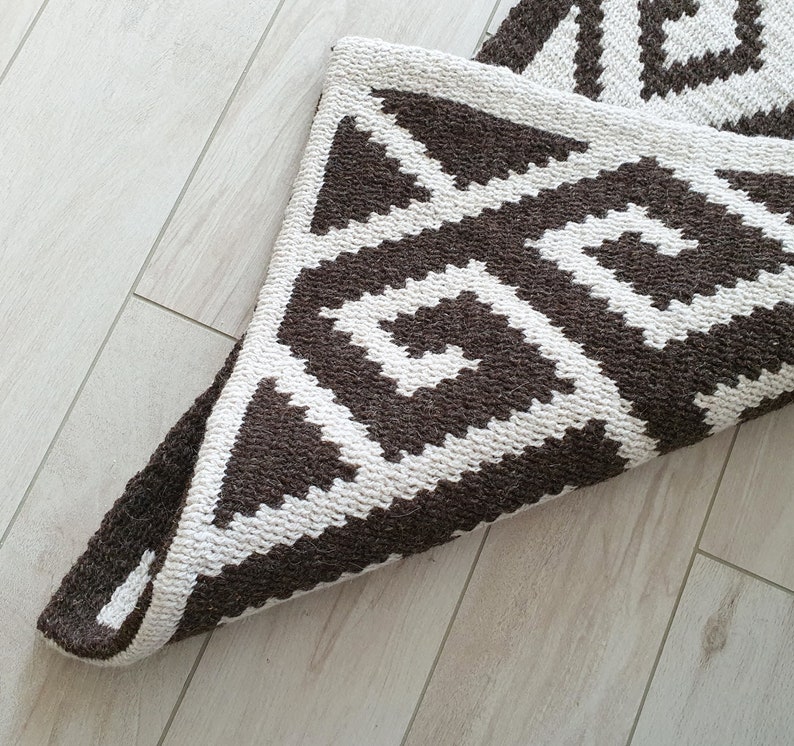 May include: A brown and white geometric patterned rug with a folded edge. The rug has a modern design with a repeating pattern of triangles and squares.