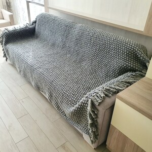 Peut inclure: Une couverture grise en tricot avec un motif textur&eacute; en forme de boules recouvre un canap&eacute;. La couverture a une frange sur les bords. Le canap&eacute; est gris clair et la pi&egrave;ce a un plancher en bois clair.