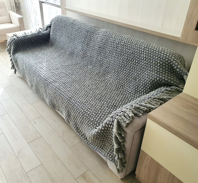 Peut inclure: Une couverture grise en tricot avec un motif textur&eacute; en forme de boules recouvre un canap&eacute;. La couverture a une frange sur les bords. Le canap&eacute; est gris clair et la pi&egrave;ce a un plancher en bois clair.