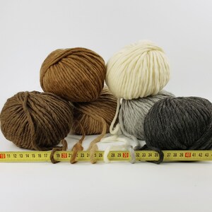 May include: Four balls of yarn in different colours: brown, beige, grey, and dark grey. The yarn is arranged in a pyramid shape with a measuring tape in front of the balls.