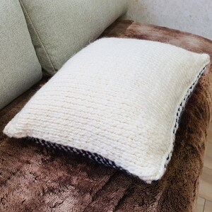 May include: A white knitted throw pillow with a black and white trim. The pillow is sitting on a brown faux fur surface.