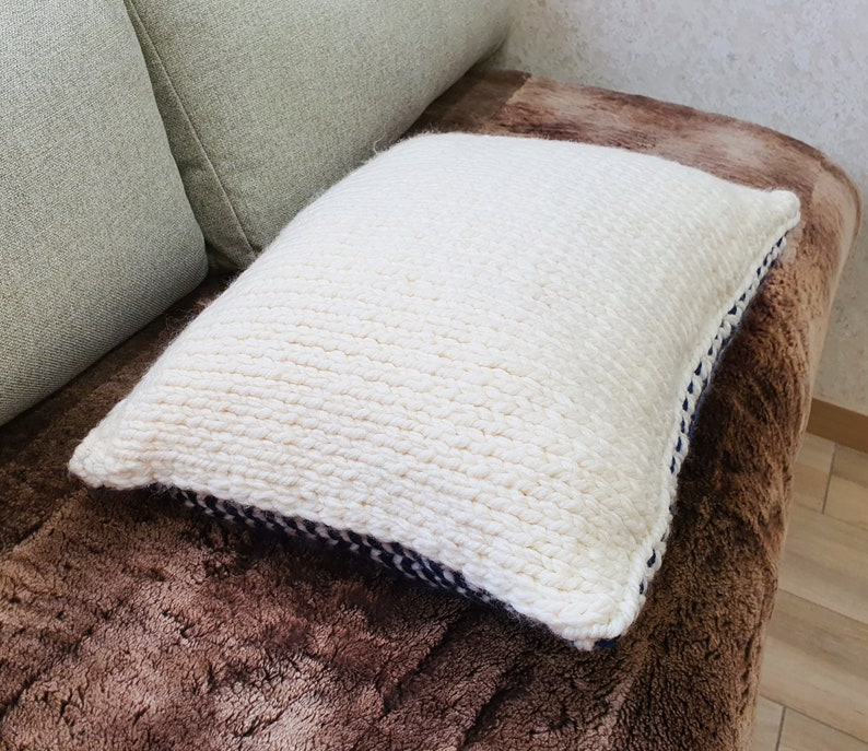 May include: A white knitted throw pillow with a black and white trim. The pillow is sitting on a brown faux fur surface.