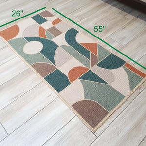 Puede incluir: Una alfombra rectangular con un patr&oacute;n geom&eacute;trico abstracto. La alfombra mide 140 cm de largo y 66 cm de ancho. Presenta formas en verde azulado, naranja, marr&oacute;n y beige. La alfombra tiene un borde beige y est&aacute; colocada sobre un suelo de madera clara.