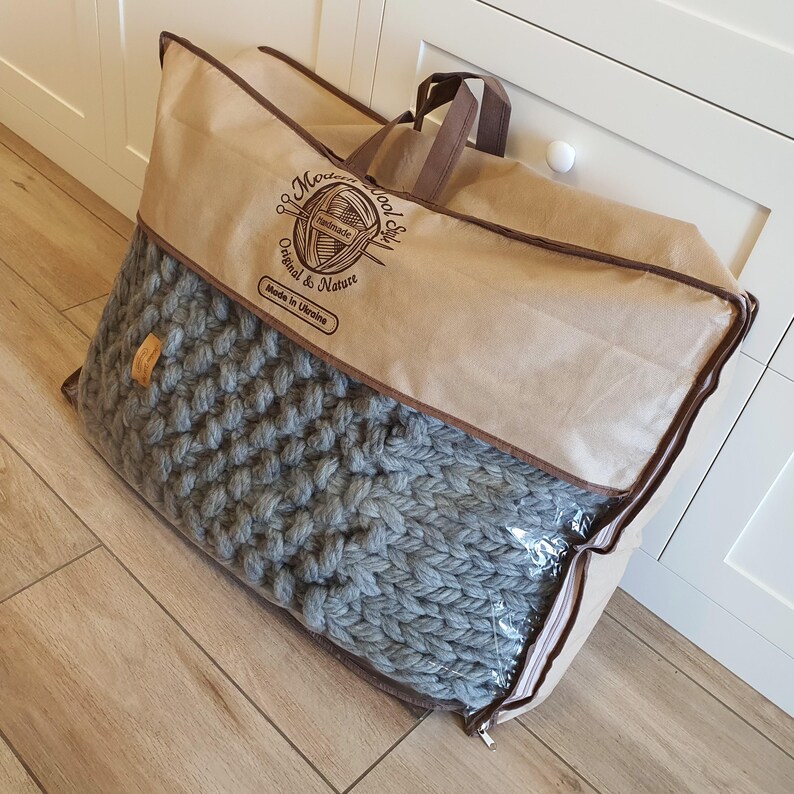 A large, gray, chunky knit blanket is packaged in a clear plastic bag inside a brown carrying case. The case has a brown zipper and handles. Text on the case reads "Modern Wool Soft Handmade Original & Nature Made in Ukraine."
