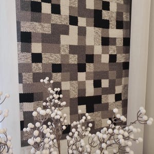 K&ouml;nnte beinhalten: Eine schwarze, wei&szlig;e und graue gewebte Wandteppich mit einem geometrischen Muster. Der Wandteppich h&auml;ngt an einer wei&szlig;en Wand.