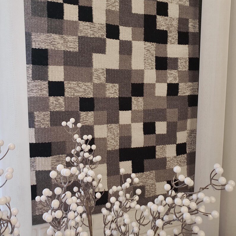 K&ouml;nnte beinhalten: Eine schwarze, wei&szlig;e und graue gewebte Wandteppich mit einem geometrischen Muster. Der Wandteppich h&auml;ngt an einer wei&szlig;en Wand.