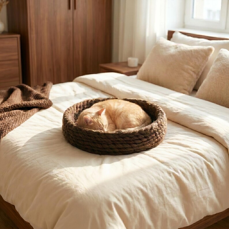 A ginger cat sleeping in a round dark brown chunky knit wool cat bed, placed on a white linen bed in a warm bedroom with walnut wood furniture, a cream pillow headboard, and soft natural window light.